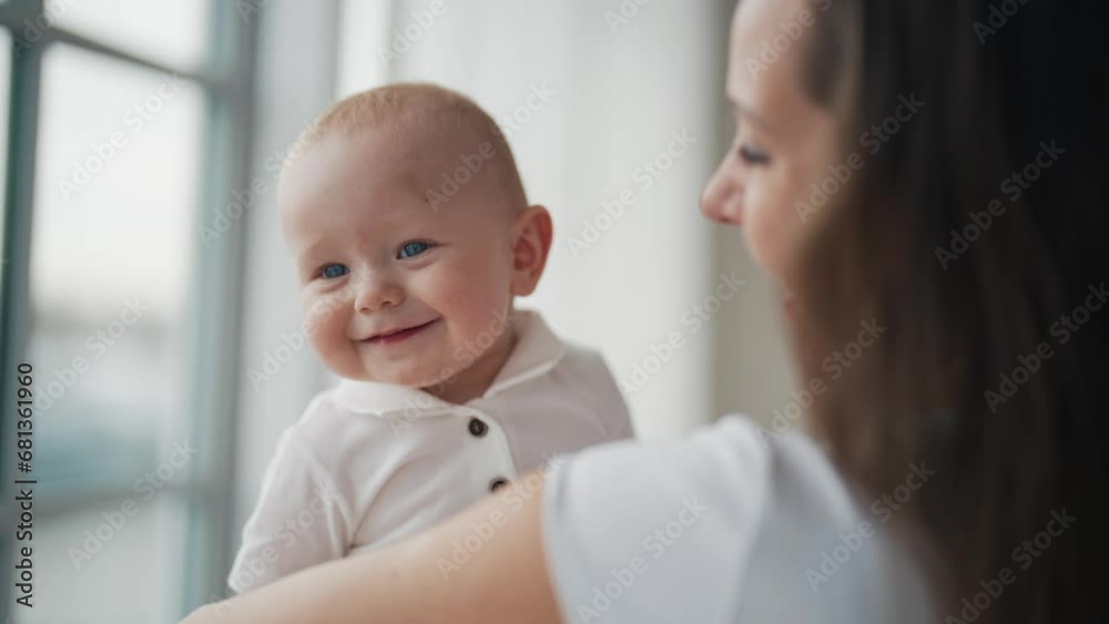 Portrait of happy family affectionate positive mom kissing baby holding in hands looking at window. Mother kisses kid at home. Motherhood, parenting, babycare, childhood, babyhood, love care concept. Portrait of happy family affectionate positive mom kissing baby holding in hands looking at window. Mother kisses kid at home. Motherhood, parenting, babycare, childhood, babyhood, love care concept.