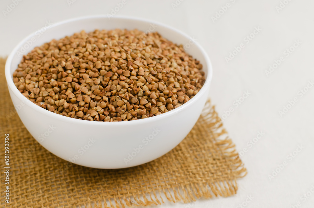 Organic dry buckwheat groats in a white bowl on a burlap napkin on a light background. Rural style. Vegetarian and vegan food. Concept of healthy eating. Horizontal orientation. Selective focus Organic dry buckwheat groats in a white bowl on a burlap napkin on a light background. Rural style. Vegetarian and vegan food. Concept of healthy eating. Horizontal orientation. Selective focus