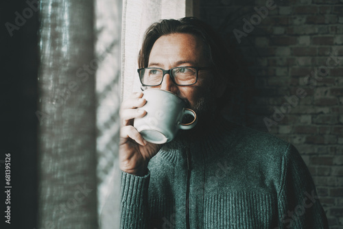 One man with thoughtful and serene expression on face drinking coffee or tea alone at home looking outside the window. Mature male people alone. Healthy mindful lifestyle concept. Wearing glasses