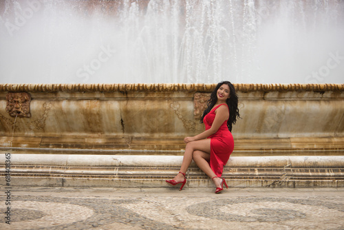 South American woman, young, beautiful, brunette, with an elegant red dress, posing sitting on a fountain in the square of Spain in Seville. Concept of beauty, fashion, ethnicity, diversity, elegance.