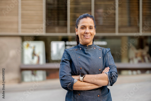 Female chef wears blue coat, broadly smiles and poses with arms crossed outdoors on the street. Restaurant worker, culinary gourmet, pastry chef.