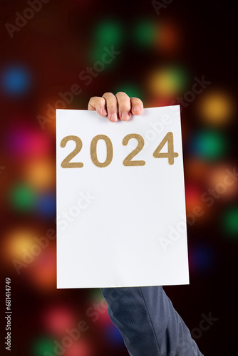 2024, Man holding cardboard with number 2024 on background of faded lights. Happy New Year