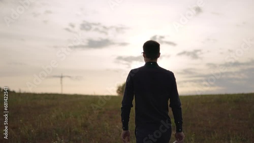 Foreman walks across field towards tower crane and rising sun on horizon. Confident man puts on protective helmet, back view. Moving towards your dreams and path to realizing your goals.
