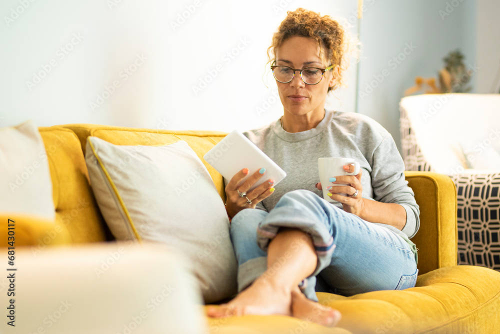 Confident caucasian adult woman sitting on the sofa reading a book using electronic device tablet reader. Pretty female people have relax and enjoy leisure activity at home laying on the couch Confident caucasian adult woman sitting on the sofa reading a book using electronic device tablet reader. Pretty female people have relax and enjoy leisure activity at home laying on the couch