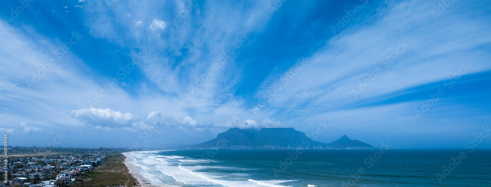 Fototapeta premium Aerial wide view of Table Mountain in Cape Town on a sunny day, clouds billowing over the mountain and beach in foregound.