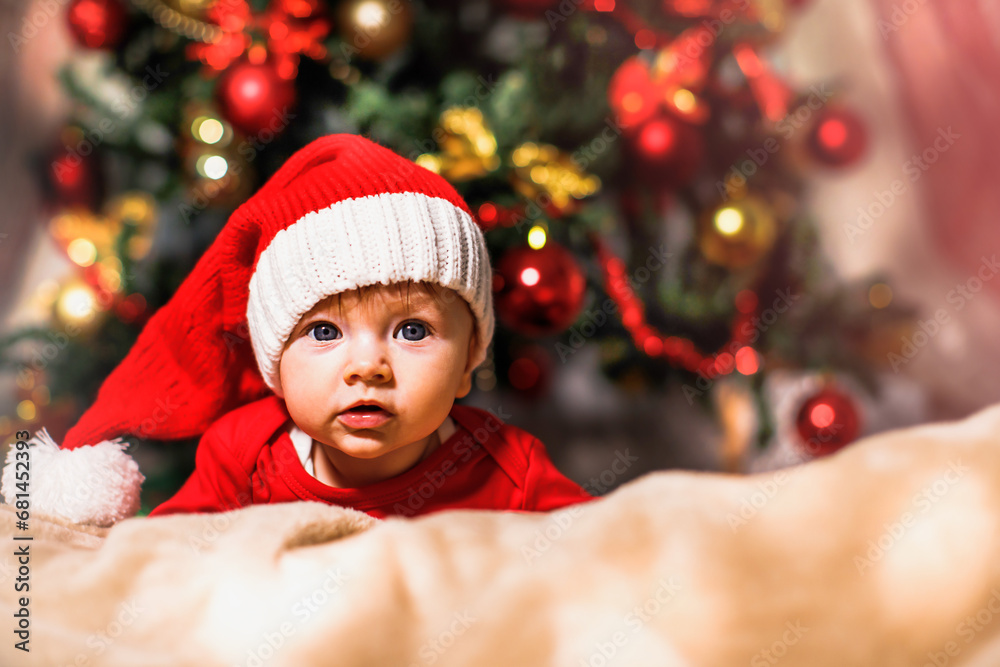 Joyful baby looking at camera in Christmas photo. Newborn baby in red cap of Santa Claus and Christmas tree in background Joyful baby looking at camera in Christmas photo. Newborn baby in red cap of Santa Claus and Christmas tree in background
