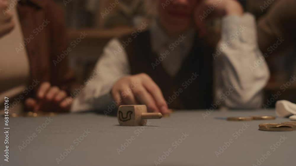 Close-up shot of hand of unrecognizable Jewish woman spinning wooden dreidel toy on table during Hanukkah festival, with visible Hebrew letters Shin - put in gelt coin, and Nun - nothing Close-up shot of hand of unrecognizable Jewish woman spinning wooden dreidel toy on table during Hanukkah festival, with visible Hebrew letters Shin - put in gelt coin, and Nun - nothing