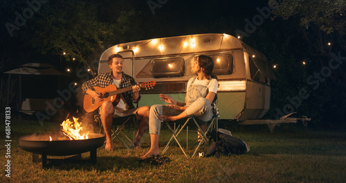 Couple in a Relationship Enjoying a Summer Evening at a Caravan Camping Area. Handsome Boyfriend Playing Acoustic Guitar Next to a Campfire, His Girlfriend is Dancing Barefoot Full of Joy