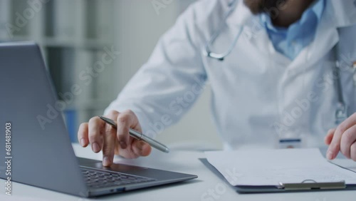 Closeup pan shot of unrecognizable male doctor looking at information in laptop and writing it down in medical history during workday in hospital photo