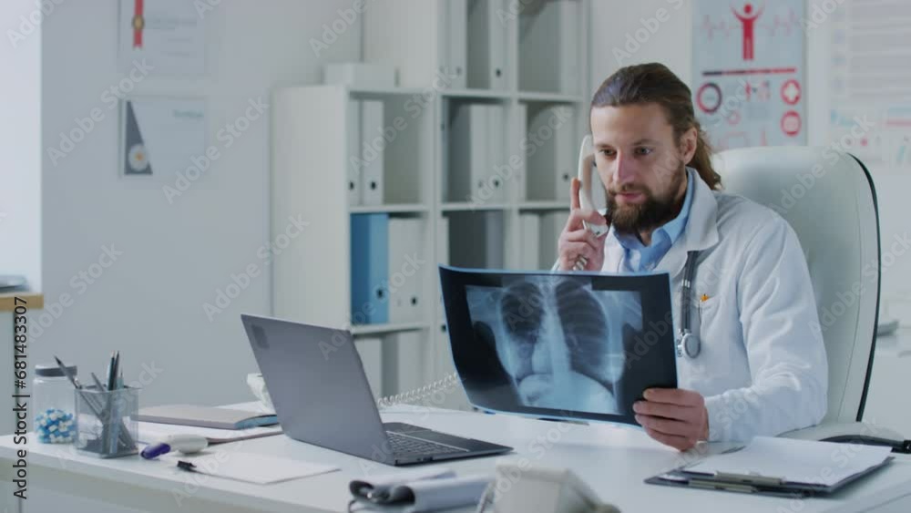Medium shot of male Caucasian practitioner speaking on phone while checking chest X-ray and typing information into laptop in medical office Medium shot of male Caucasian practitioner speaking on phone while checking chest X-ray and typing information into laptop in medical office