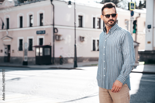 Portrait of handsome confident stylish hipster lambersexual model. Sexy modern man dressed in blue shirt and trousers. Fashion male posing on street background in Europe city at sunset. In sunglasses