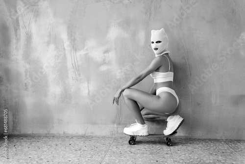 Beautiful sexy woman in underwear. Model wearing bandit balaclava mask. Hot seductive female in lingerie posing near grey wall in studio. Crime and violence. Sits at penny skateboard. Black and white