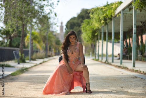 Young, Hispanic, beautiful, brunette, young woman in an elegant salmon-colored dress, posing independent and empowered, sitting on a chair outdoors. Concept of beauty, fashion, trend, elegance.