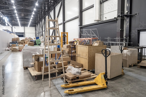 Hangar with boxes and stepladder. Industrial premises with pallet jack near containers. Factory hangar with boxes filled with building materials. Industrial warehouse. Mess in storage space photo