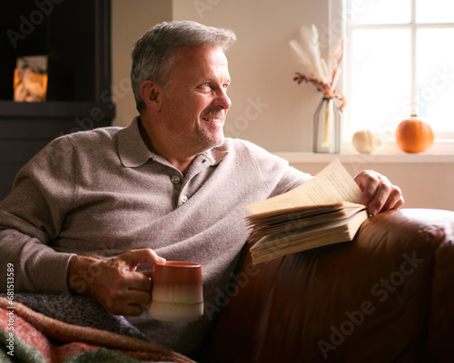 Mature Man At Home In Winter Jumper On Sofa With Warming Hot Drink Of Coffee In Mug Reading Book photo