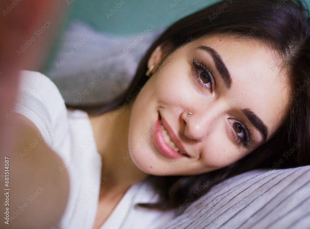 Beautiful brunette caucasian young woman taking selfie lying on her bed at home, smiling face expression closeup portrait photography