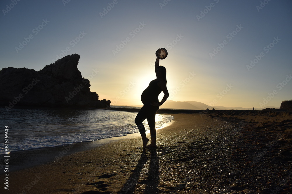 pregnant woman in profile on the beach with backlit sunset and a hat