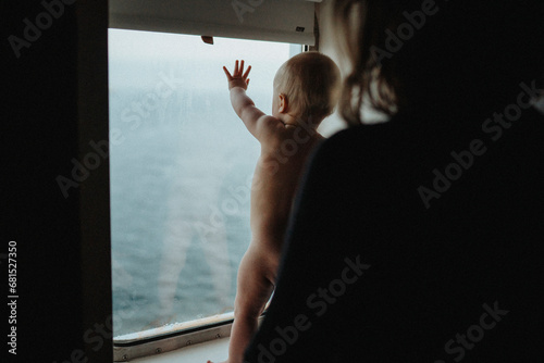 Mother supporting naked baby who is standing on window sill photo