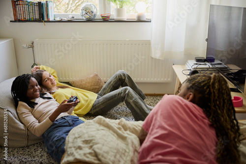 Smiling teenage girl holding game controller while lying down with female friends at home photo
