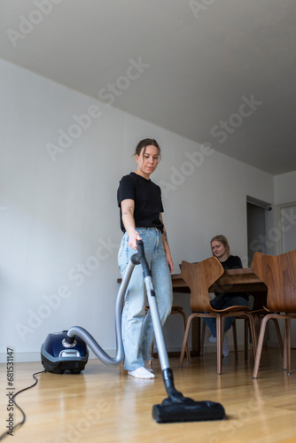 Young woman cleaning hardwood floor with vacuum cleaner near girlfriend using laptop at home photo