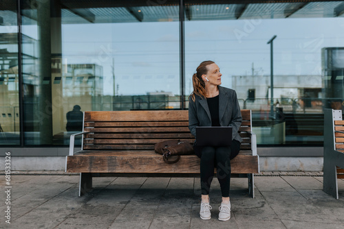 Full length of businesswoman with laptop sitting on bench while waiting at railroad station photo
