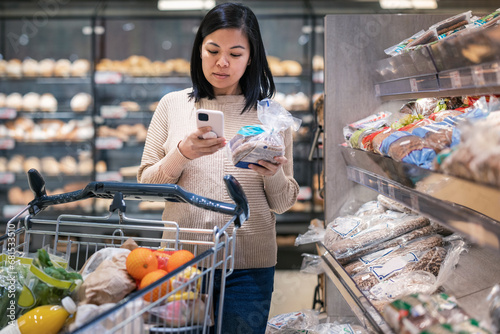 Young woman using smart phone while holding bread package and shopping in supermarket