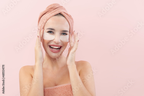 Skincare. Very happy young woman laughing, applying cosmetic eye patches mask, reduces wrinkles, wears wrapped towel on head, isolated on pink background. Facial treatment, beauty and spa concept.