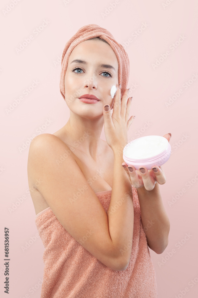Skincare, pretty young woman applying moisturizing cream on face, lotion or mask for skin lifting and anti-aging, wears wrapped towel on the head and holds the jar in hand. Isolated On pink background Skincare, pretty young woman applying moisturizing cream on face, lotion or mask for skin lifting and anti-aging, wears wrapped towel on the head and holds the jar in hand. Isolated On pink background
