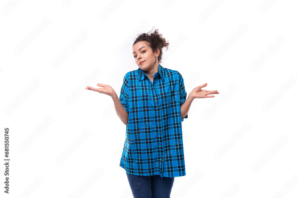 young curly brunette woman in a blue plaid shirt doubts and gesticulates against the background with copy space