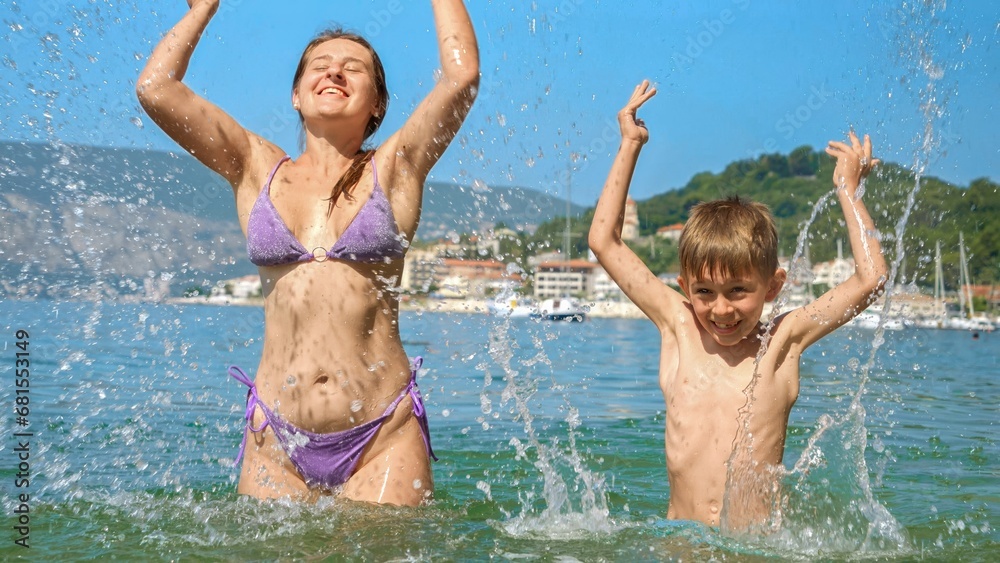 Happy cheerful excited boy splashing water with mother while relaxing on sunny sea beach