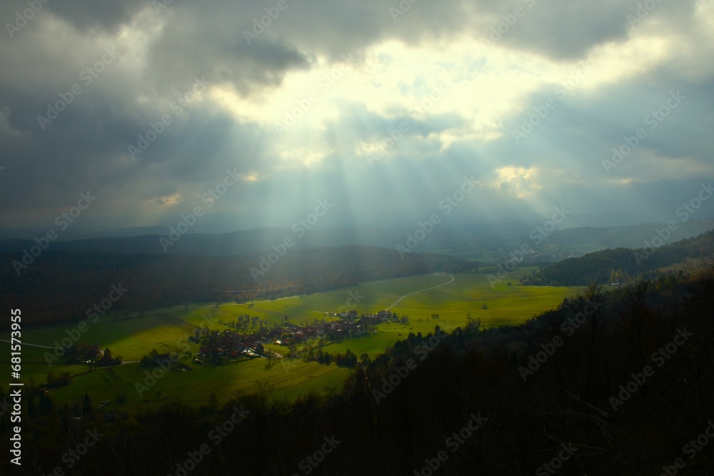 Obraz premium View of Strmca village at a field surrounded by forest on a cloudy day lit by sunlight in Notranjska, Slovenia