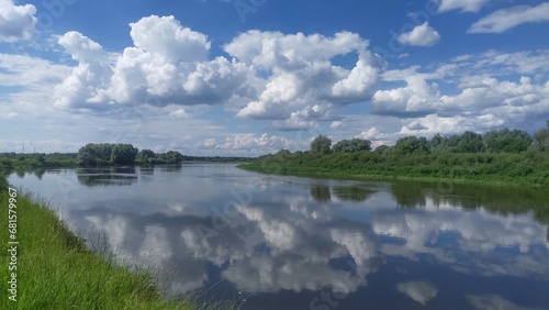 There are bushes and willows growing on the grassy banks of the river. On the water - ripples and reflection of trees, blue sky and clouds. On the far bank is a forest. Sunny summer weather