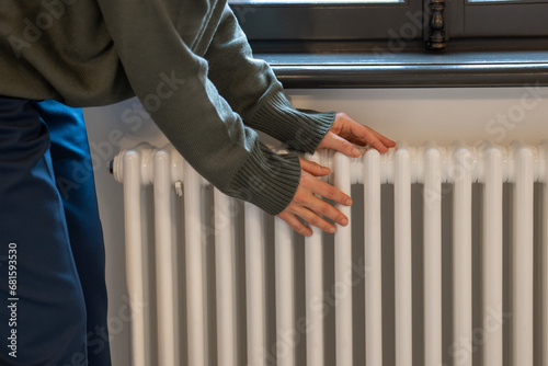 Woman warming hands near radiator at home after walking in cold winter weather, female touching barely warm battery during heating season, person near window checking heating system photo