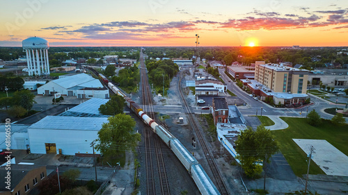 Golden sun setting on horizon with hotel and water tower split by train on railroad tracks aerial