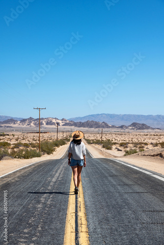 Young woman with hat and wearing shorts and a shirt walking in the middle of an asphalt road in Mojave Desert  California