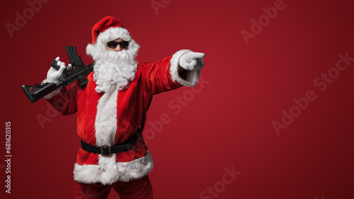 A man dressed as Santa Claus, wearing sleek black sunglasses, poses with toy machine guns against a bold red backdrop