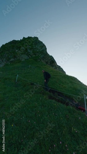 Vertical video. Male backpacker climbing stairs rope summer morning mountain summit sunny ski resort aerial view. Travel man hiker going upstairs natural cliff rock covered green grass effort journey photo