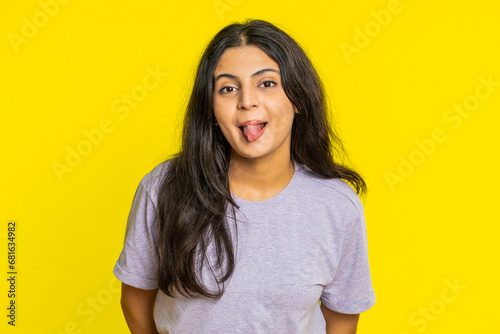 Funny comical playful Indian young woman making silly facial expressions grimacing fooling around, showing tongue, idiotic expression. Comedian Arabian girl on yellow background. People lifestyles photo