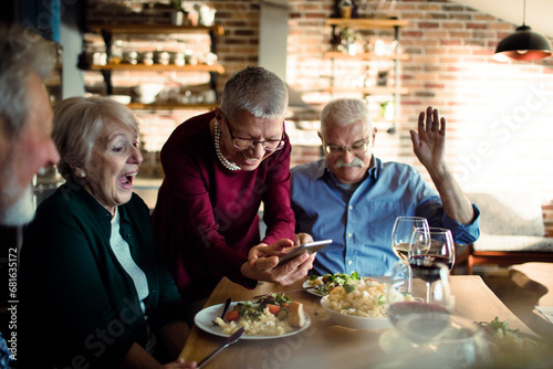 Senior woman showing something funny on smartphone during lunch with friends at home