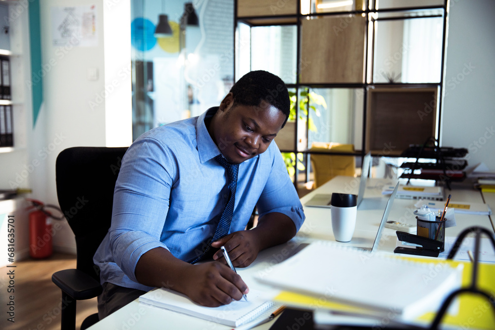 Black businessman writing notes in notebook sitting at workplace in office