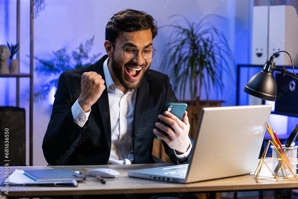 Happy Indian businessman freelancer use mobile smartphone celebrating success victory, winning lottery jackpot goal achievement, play game, good positive news, triumph working at home office workplace Happy Indian businessman freelancer use mobile smartphone celebrating success victory, winning lottery jackpot goal achievement, play game, good positive news, triumph working at home office workplace