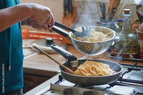 Cooking traditional recipes at home. Unrecognizable woman adds spaghetti from a wringer to a pan on the kitchen stove to mix with bolognese sauce. photo