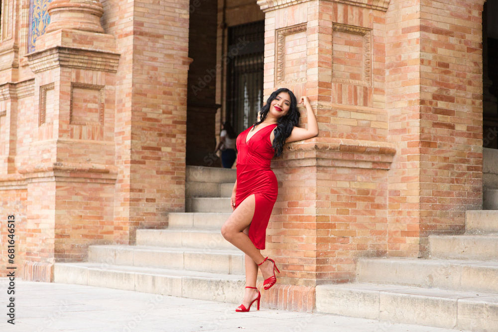 Young and beautiful brunette and latin woman, dressed in short red dress and red shoes visits the famous square in seville, spain. The woman is leaning on a brick column and is happy. Travel concept.