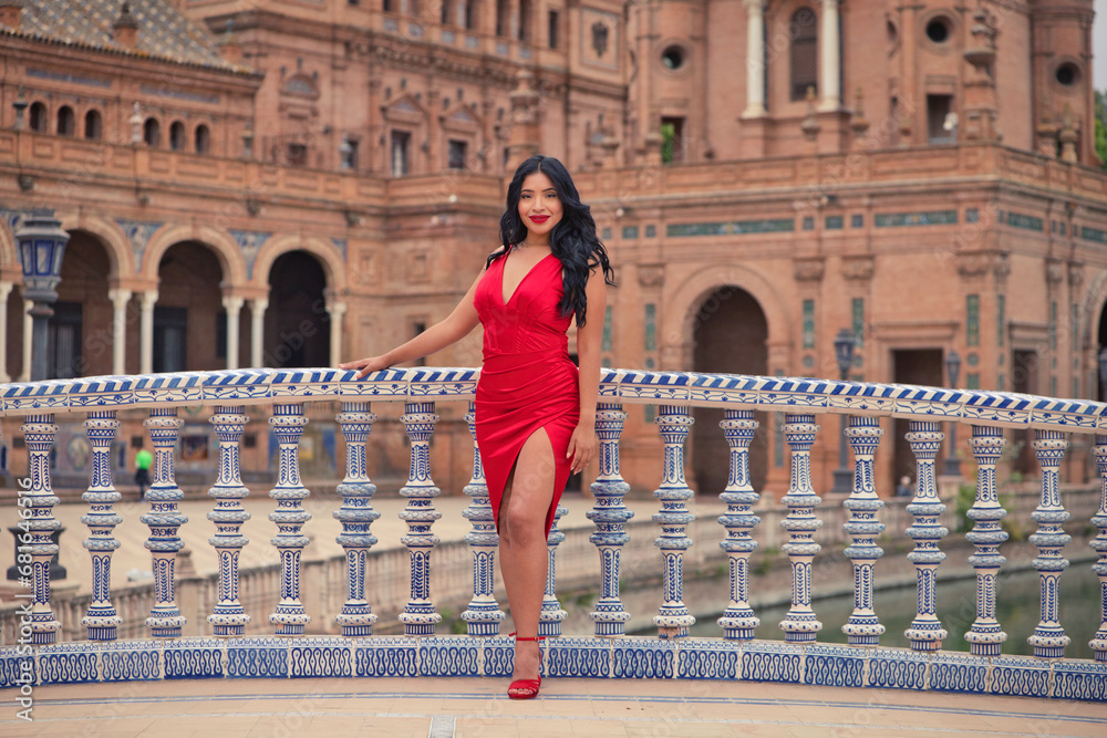 Young and beautiful brunette and latin woman, dressed in short red dress and red shoes visits the famous square in seville, spain. Woman leaning on the railing of a tiled bridge and is happy. Travel.