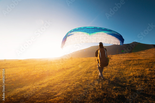male paraglider takes off from a yellow field with a blue parachute against the backdrop of hills and small mountains. Paragliding