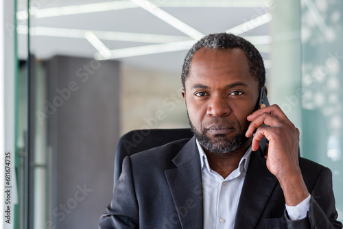 Close-up portrait of a senior African-American man sitting at a desk in the office, talking on the phone and looking seriously at the camera.