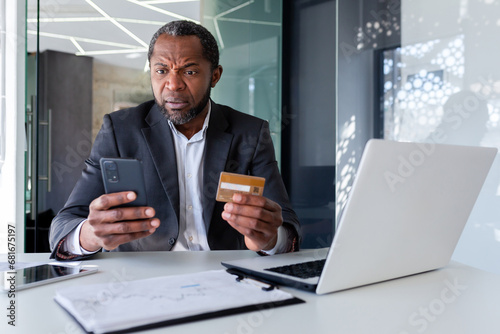 Worried and upset senior african american man working in office, holding phone and credit card.