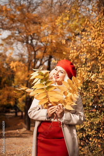 autumn woman in a red beret  a light coat and a red skirt  against the backdrop of an autumn park with yellow leaves in her hands