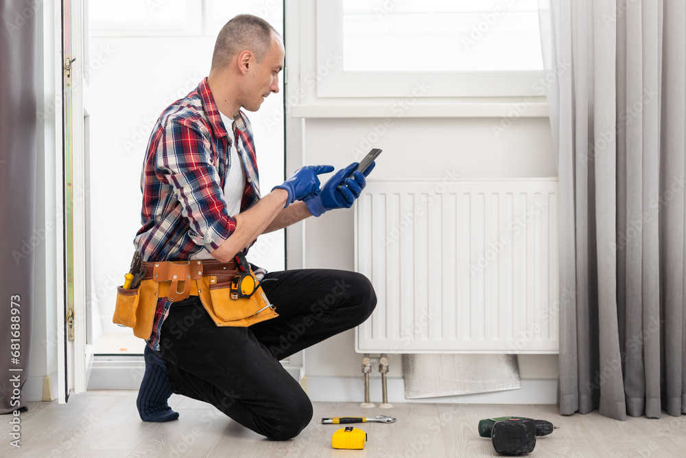 Man in work overalls using wrench while installing heating radiator in room. Young plumber installing heating system in apartment. Concept of radiator installation, plumbing works and home renovation Man in work overalls using wrench while installing heating radiator in room. Young plumber installing heating system in apartment. Concept of radiator installation, plumbing works and home renovation