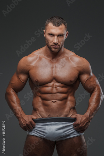 Portrait of a rugged man with a well-groomed model appearance, wearing white briefs, and showcasing his bare muscular torso against a gray background © Fxquadro